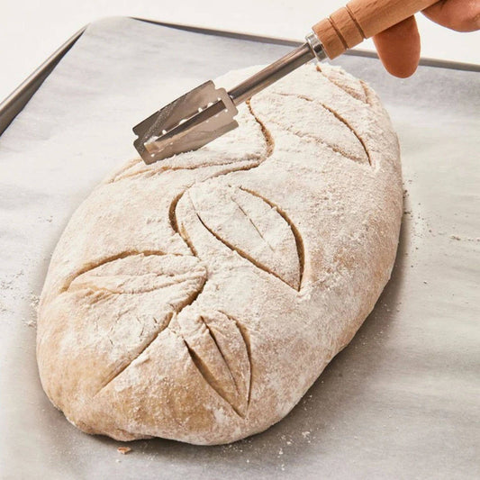 Loaf of bread with a decorative cut pattern being scored with a knife on a baking sheet.