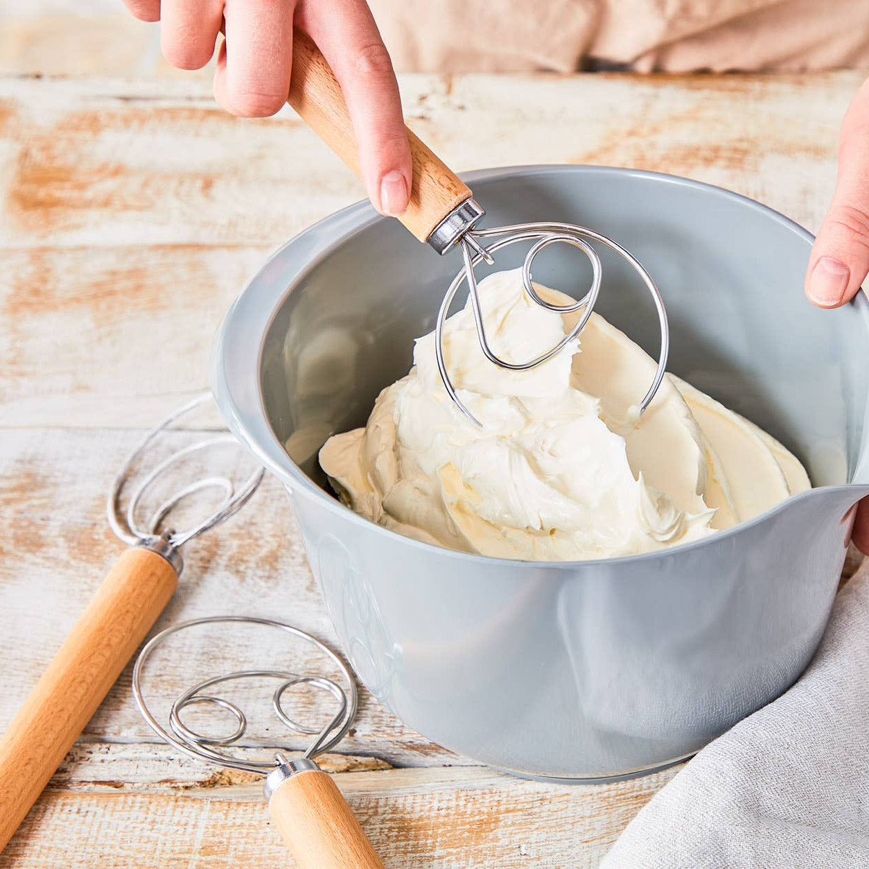 Person whisking cream in a grey bowl on a wooden surface using a danish dough whisk