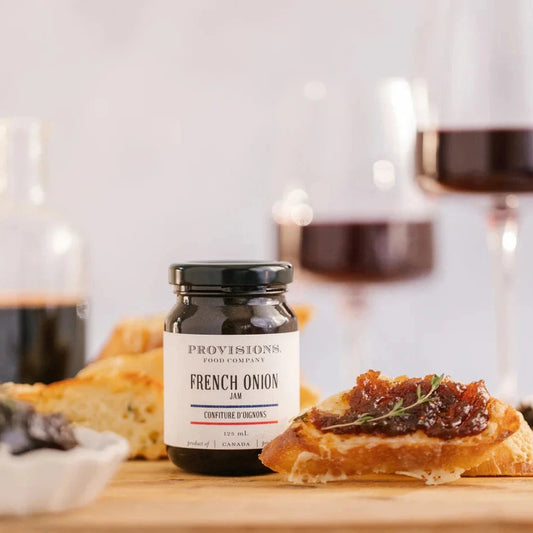 A jar of French Onion Jam on a wooden table accompanied by slices of bread and a glass of red wine in the background.