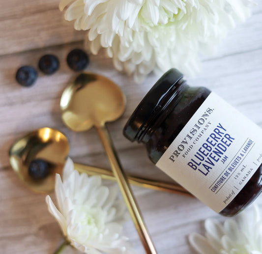 A glass jar labeled 'Blueberry Lavender' jam, alongside a golden spoon and fresh blueberries on a wooden surface with white flowers in the background.