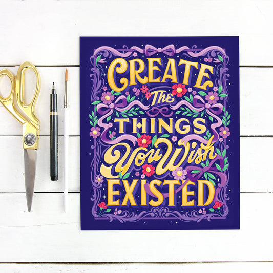 Motivational poster with 'Create the things you wish existed' text on a white wooden surface with scissors and a pen.