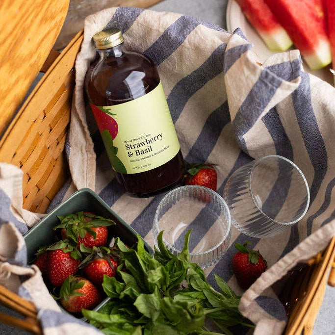 Strawberry & Basil syrup bottle on a picnic blanket with strawberries, watermelon, and flowers.