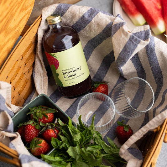 Strawberry & Basil syrup bottle on a picnic blanket with strawberries, watermelon, and flowers.