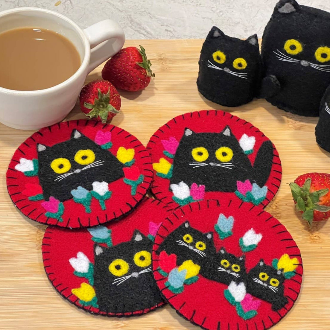 Cat-themed coasters with black cats on red backgrounds, placed on a wooden surface with a cup of coffee and strawberries.