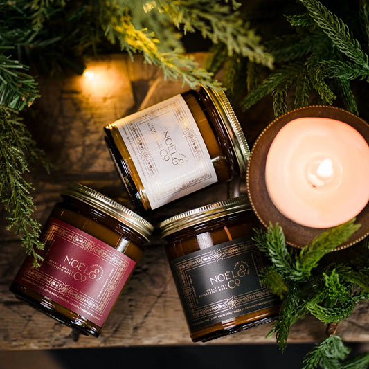 Jars of Noel & Co. Christmas candles with a lit candle on a wooden surface surrounded by greenery.