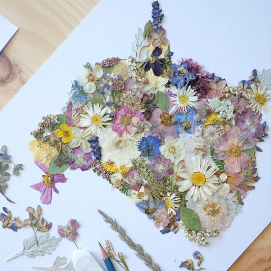 Pressed flowers and leaves arranged in the shape of New Brunswick, Canada on a piece of paper with a pencil and another leaf on a wooden surface.
